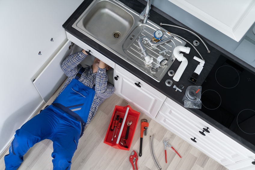 Plumber Examining Kitchen Pipes Person in blue overalls repairing pipes under a kitchen sink with tools and parts spread out on the floor.