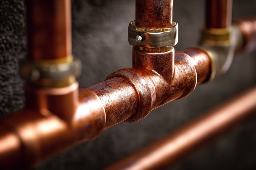 Close-up of copper plumbing pipes with a T-joint and metal clamp against a dark background.