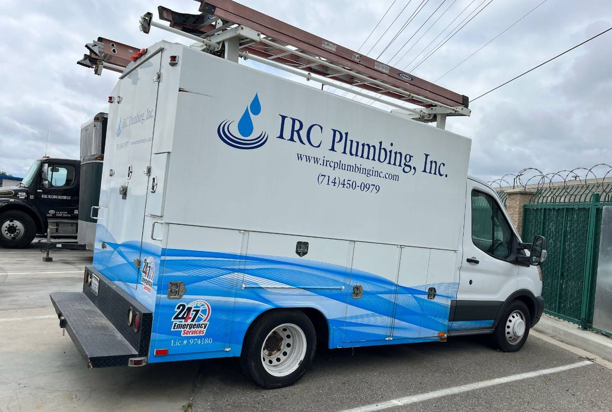 IRC Plumbing, Inc. A white IRC Plumbing, Inc. service van with a ladder on top is parked in a lot near a fence.
